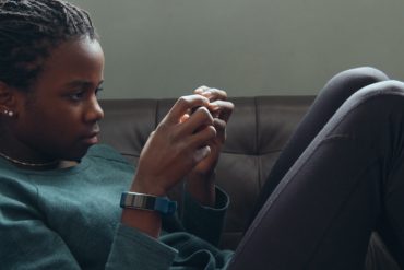 A young black girl laying on a couch, playing a game on her phone.